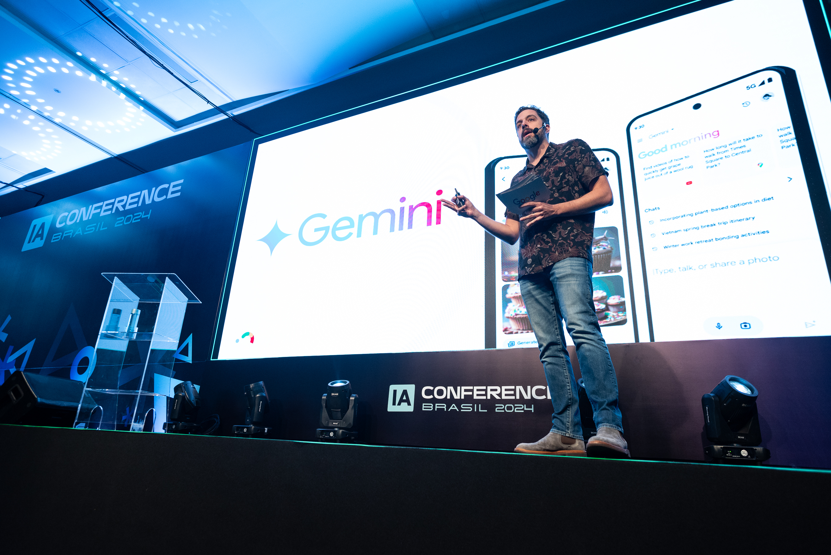 A imagem é uma fotografia de Alex Freire no palco do evento: um homem branco, de cabelo e barba escuros, de calça jeans, bota marrom e camisa de mangas curtas florida preta e marrom