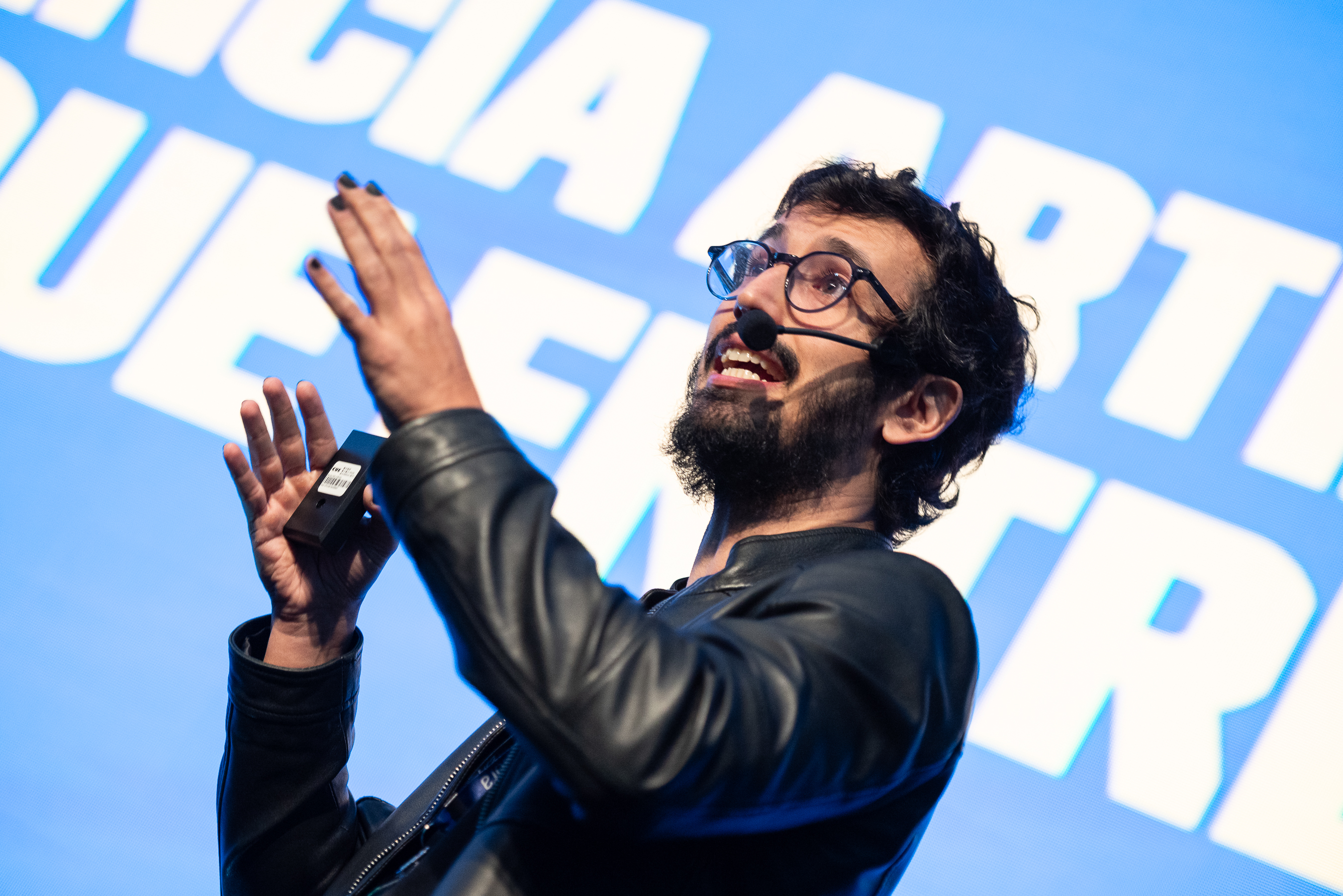 A imagem é uma fotografia de Paulo Silveira no palco do evento: um homem branco, de cabelos e barbas escuras, de camiseta preta e jaqueta de couro preta