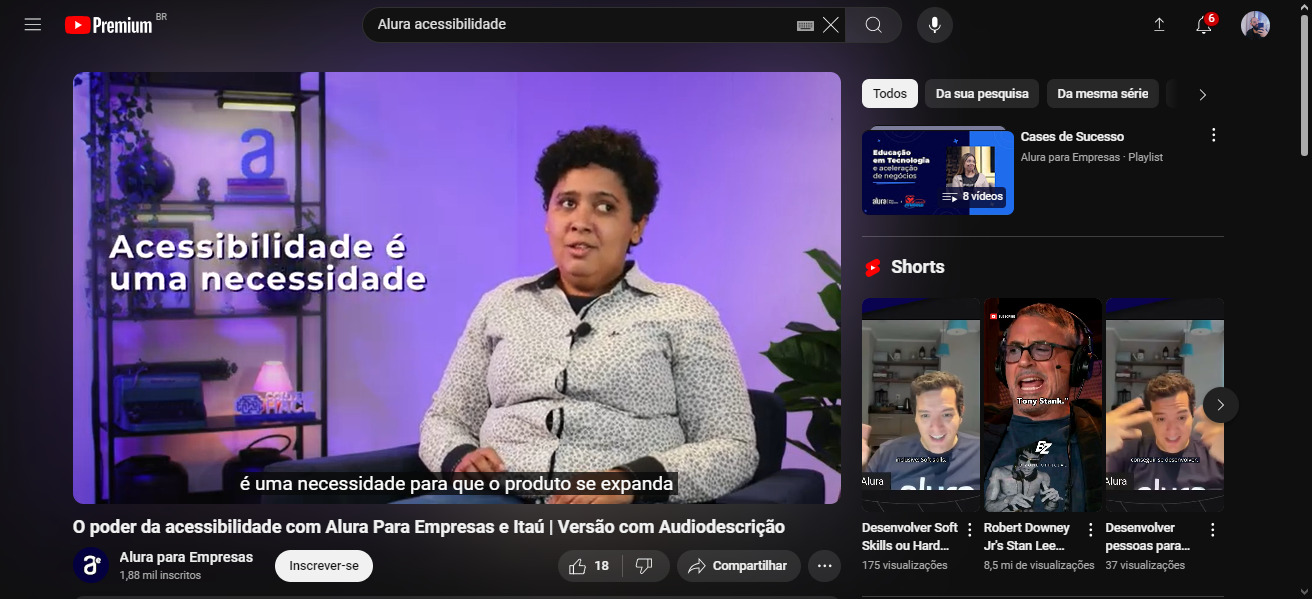 Vídeo no canal da Alura pelo YouTube chamado “O poder da acessibilidade com Alura Para Empresas e Itaú | Versão com Audiodescrição”. No vídeo aparece uma mulher de cabelo curto e blusa branca de manga longa, sentada numa poltrona. No próprio vídeo está aparecendo um texto escrito “Acessibilidade é uma necessidade”, e na parte inferior a legenda do YouTube em tempo real com a fala.