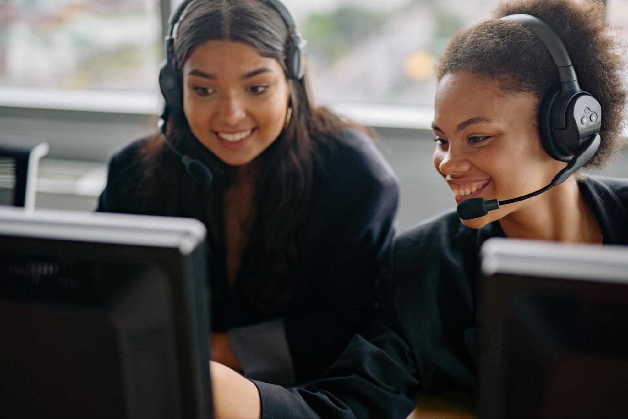 Duas mulheres com headset sorrindo e olhando para a tela do computador.