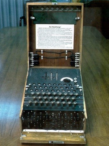 Imagem que mostra a máquina Enigma. Uma máquina eletromecânica envolvida por um caixote de madeira similar a uma mala. A máquina está aberta, seu interior é preto e apresenta um teclado com letras, acima do teclado há alguns sinalizadores das letras que acendiam após as teclas serem selecionadas. Abaixo do teclado há os rotores à mostra. Na tampa aberta há uma mensagem colada na caixa.