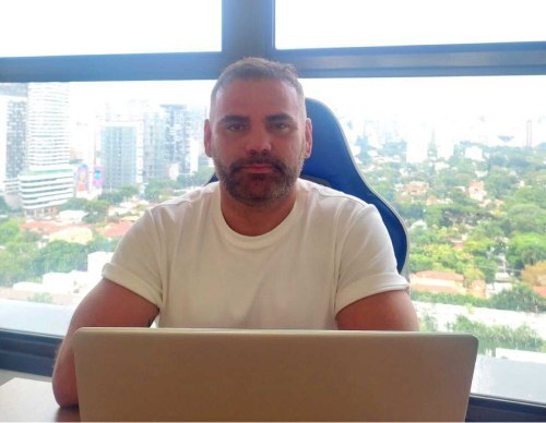 Homem branco com barba e cabelos castanhos. Veste uma camisa branca e a sua frente há um notebook aberto. Ao fundo há uma janela de vidro que mostra uma visão panorâmica de uma cidade com prédios e árvores.