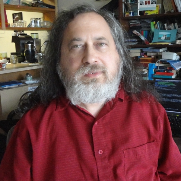 Foto retrato de um homem branco de cabelos grisalhos, barba branca e olhos verdes, usando uma camisa vermelha. Ao fundo existe um prateleira cheia de livros e eletrodomésticos