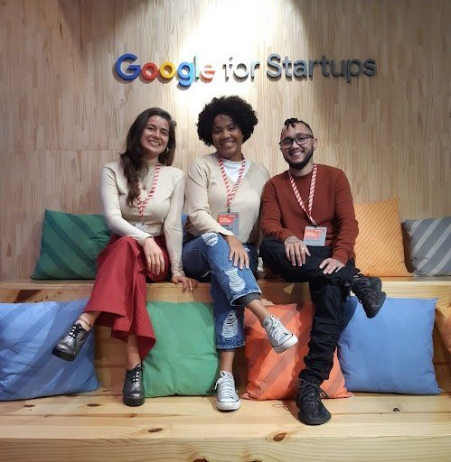 Foto com três pessoas, duas mulheres e um homem, sentados em um banco com almofadas. Na parede de fundo, escrito "Google for Startups"