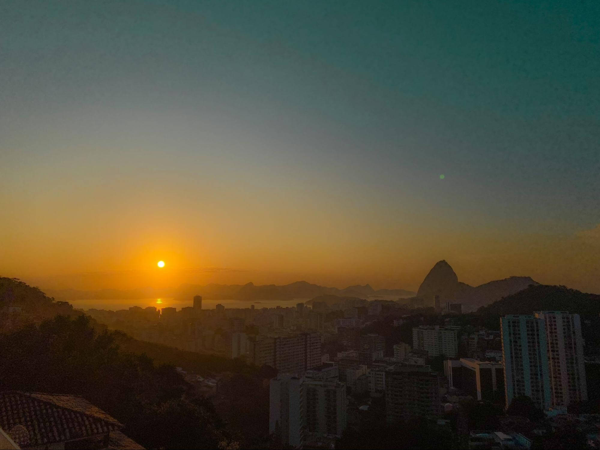 Duas imagens de um por do sol visto de cima da cidade, aparecendo vários prédios e casas. na primeira com luz natural e na segunda mais escurecida, quase não percebemos direito os prédios.