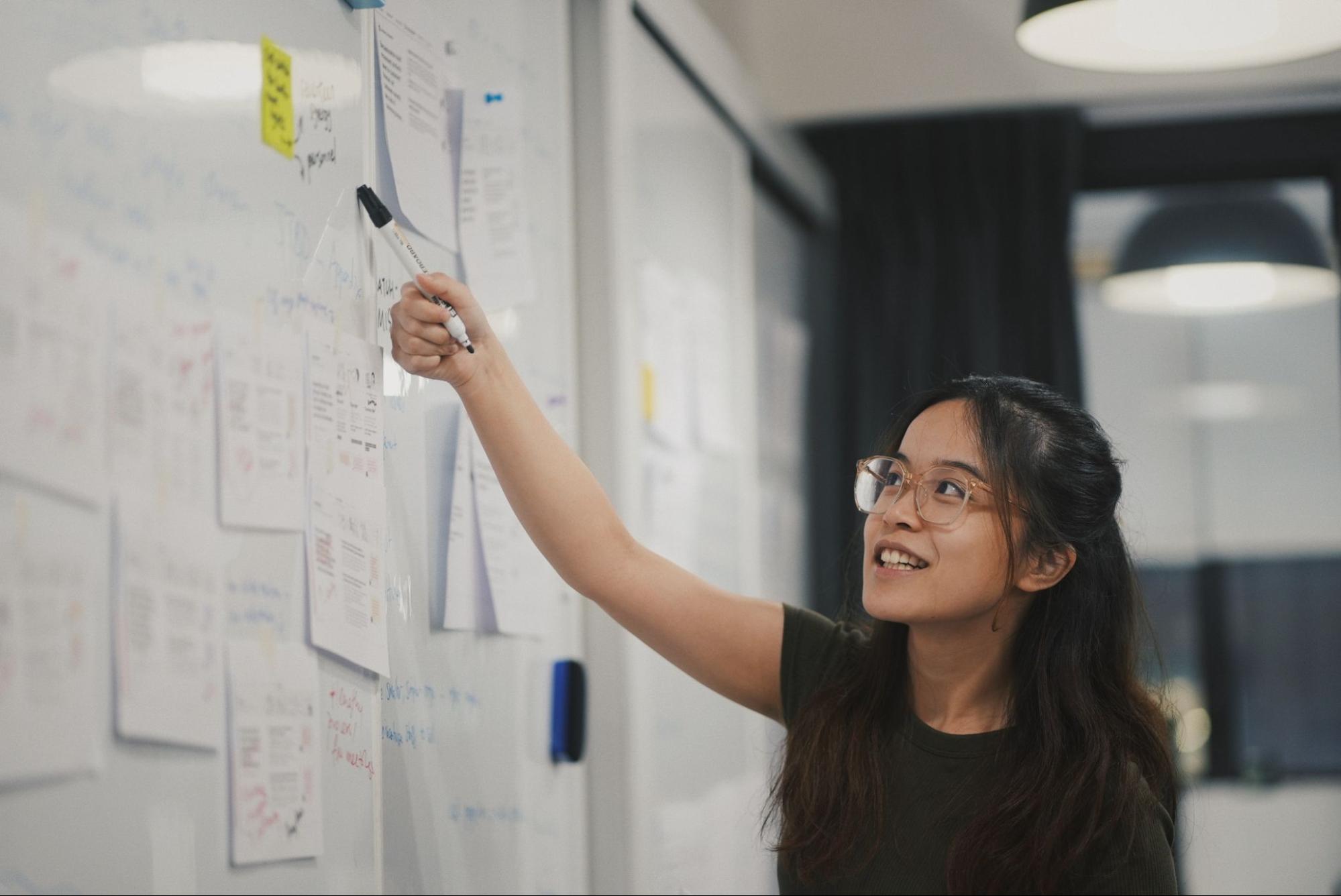 alt text: foto de uma mulher em pé, olhando para um quadro branco cheio de papéis pendurados e apontando para dois post-its específicos