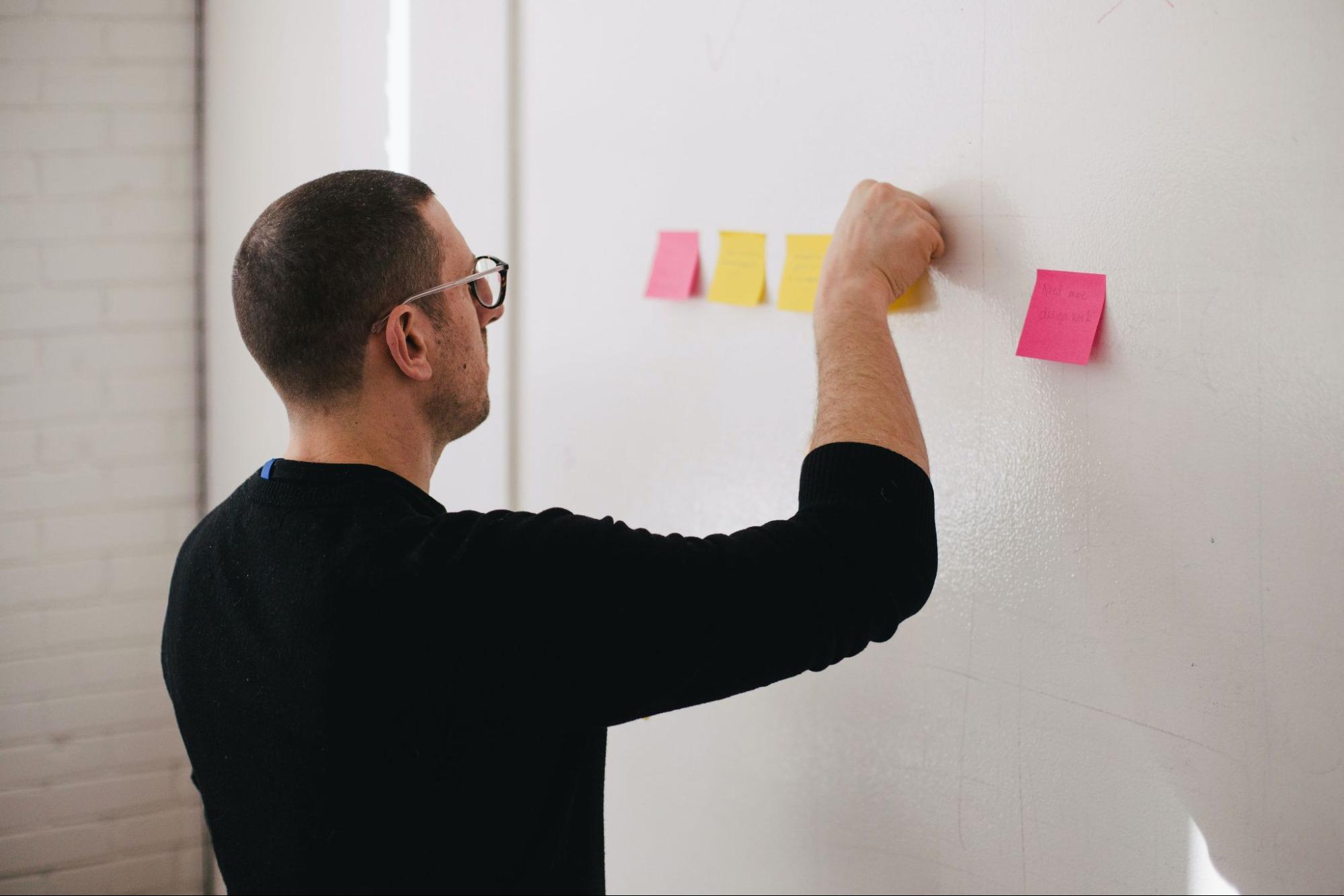 alt text: homem em pé colando alguns post-its coloridos na lousa branca pendurada na parede