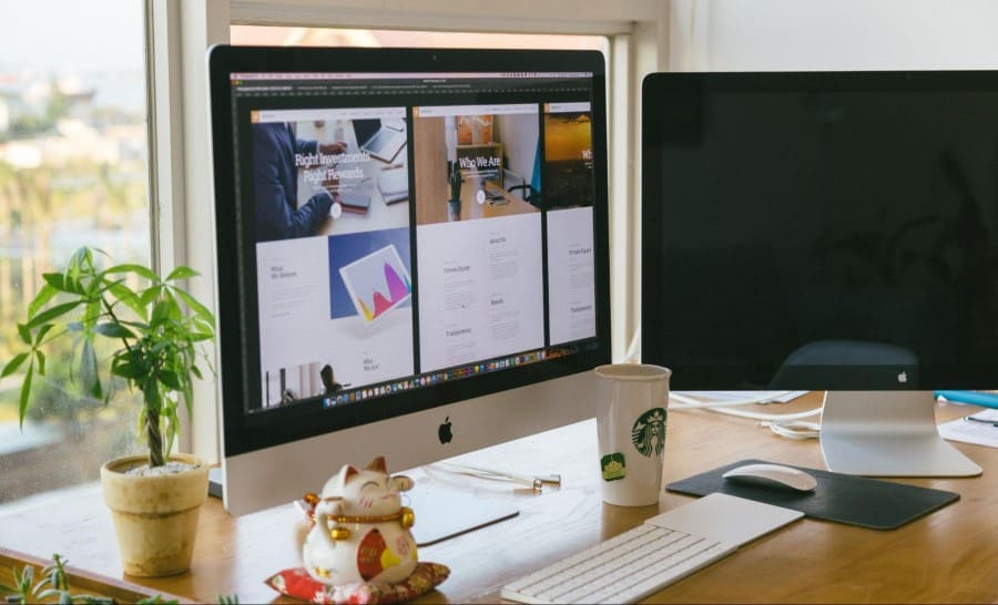 Imagem contendo dois monitores em cima de uma mesa. Um dos monitores está desligado e o outro está ligado com um projeto de interface aberto. Na mesa, no canto esquerdo há um pequeno vaso com uma planta, uma porcelana de gatinho. Já no centro da mesa, há um copo, um teclado e um mouse em cima de um mousepad preto.