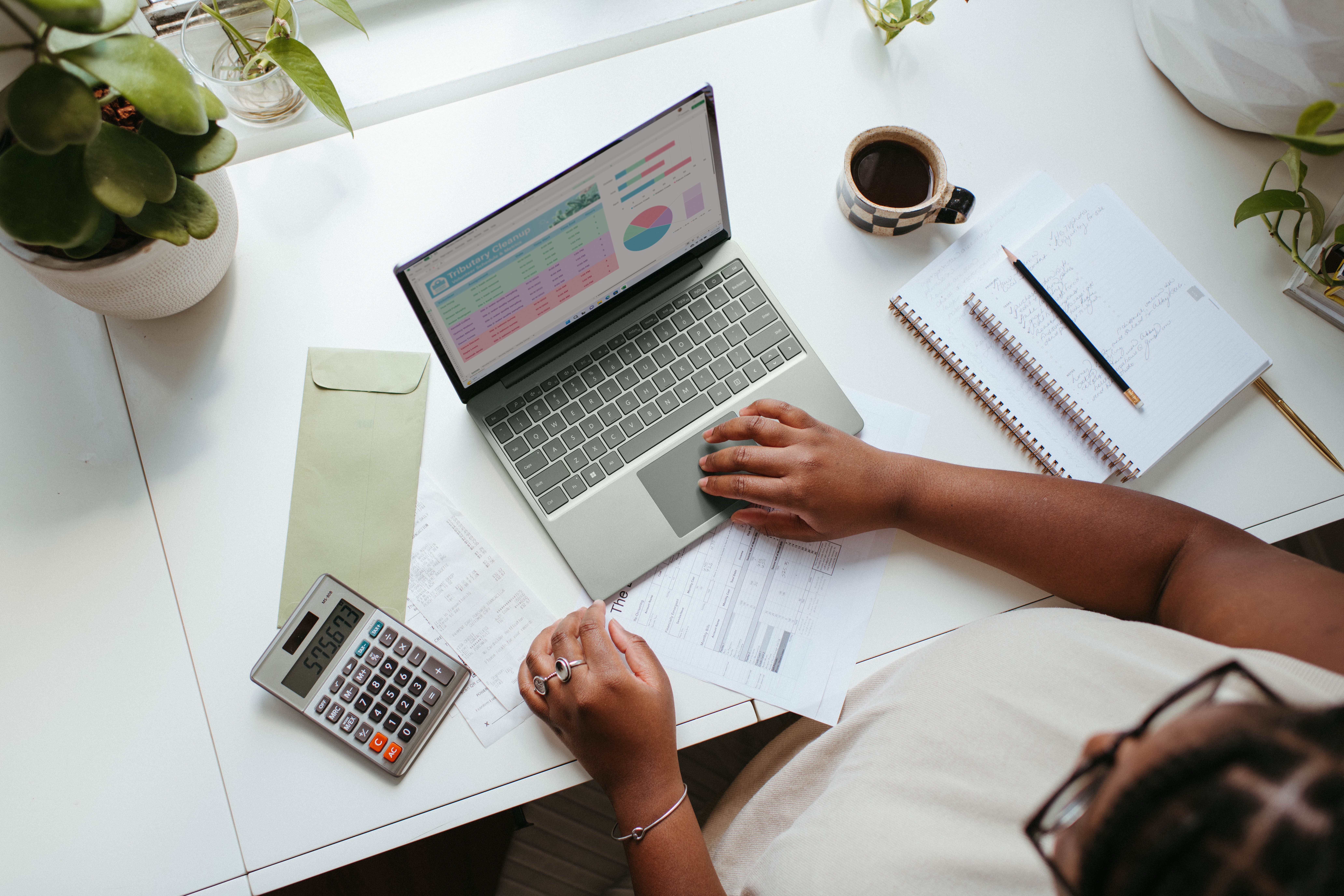 Imagem de uma mulher sentada de frente para uma mesa e utilizando um computador e com alguns objetos à sua volta – dois cadernos com um lápis sobre eles, alguns papéis, uma calculadora, um envelope, uma xícara com café e algumas plantas.