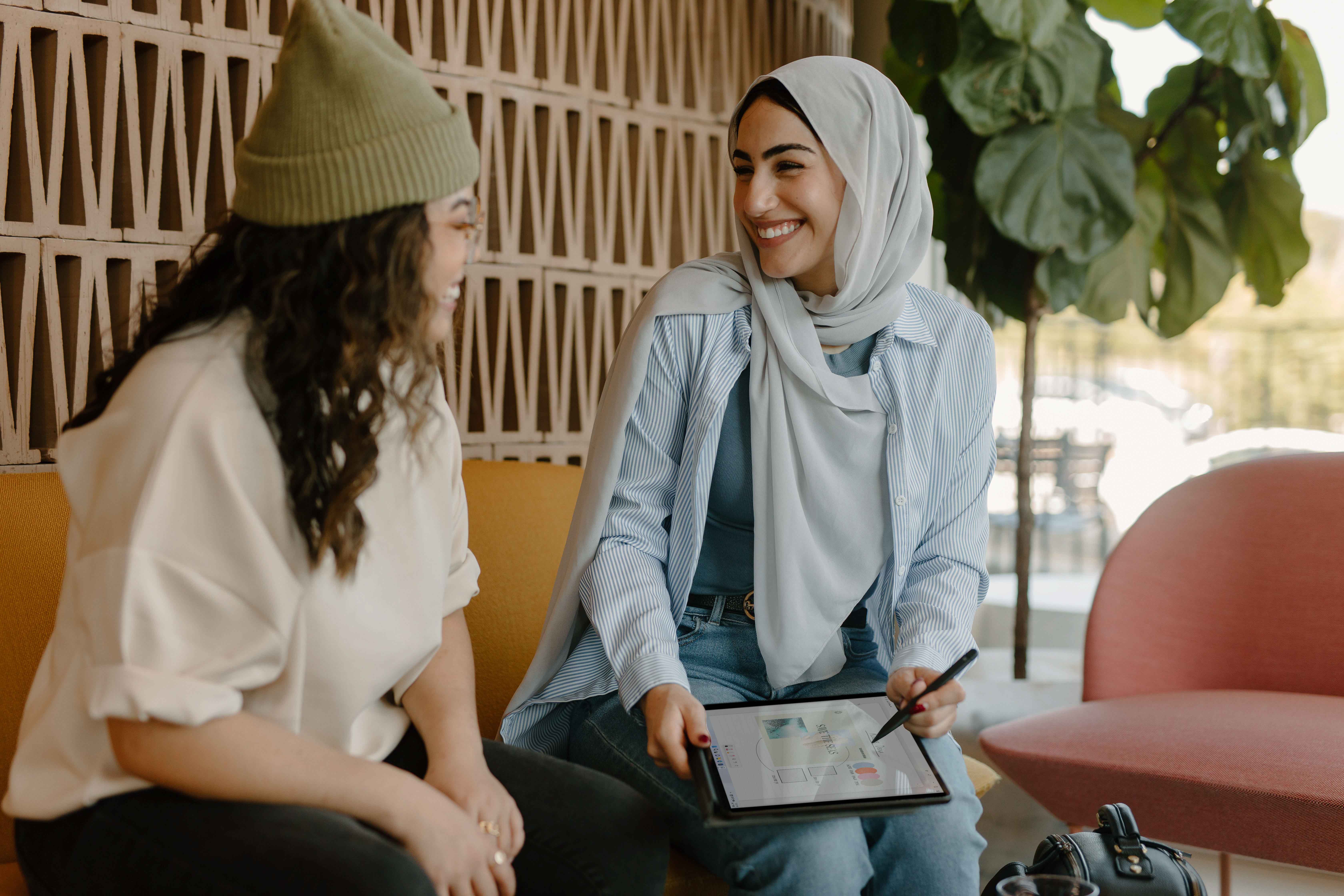 Imagem de duas mulheres sentadas em um sofá e conversando. Uma delas segura um tablet na mão direita e uma caneta na mão esquerda.