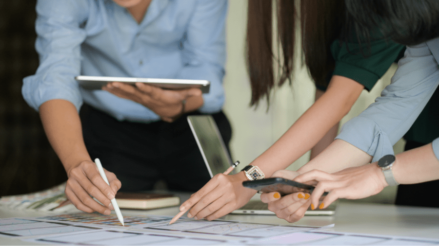 Imagem de uma mesa na parte inferior com papéis em cima dela com informações que estão borradas, além de um caderno fechado e um notebook aberto, tendo três pessoas ao redor dessa mesa, interagindo com os papéis enquanto a primeira pessoa segura um tablet e uma caneta, a segunda pessoa tem uma caneta na mão esquerda e a terceira pessoa segura um celular com ambas as mãos