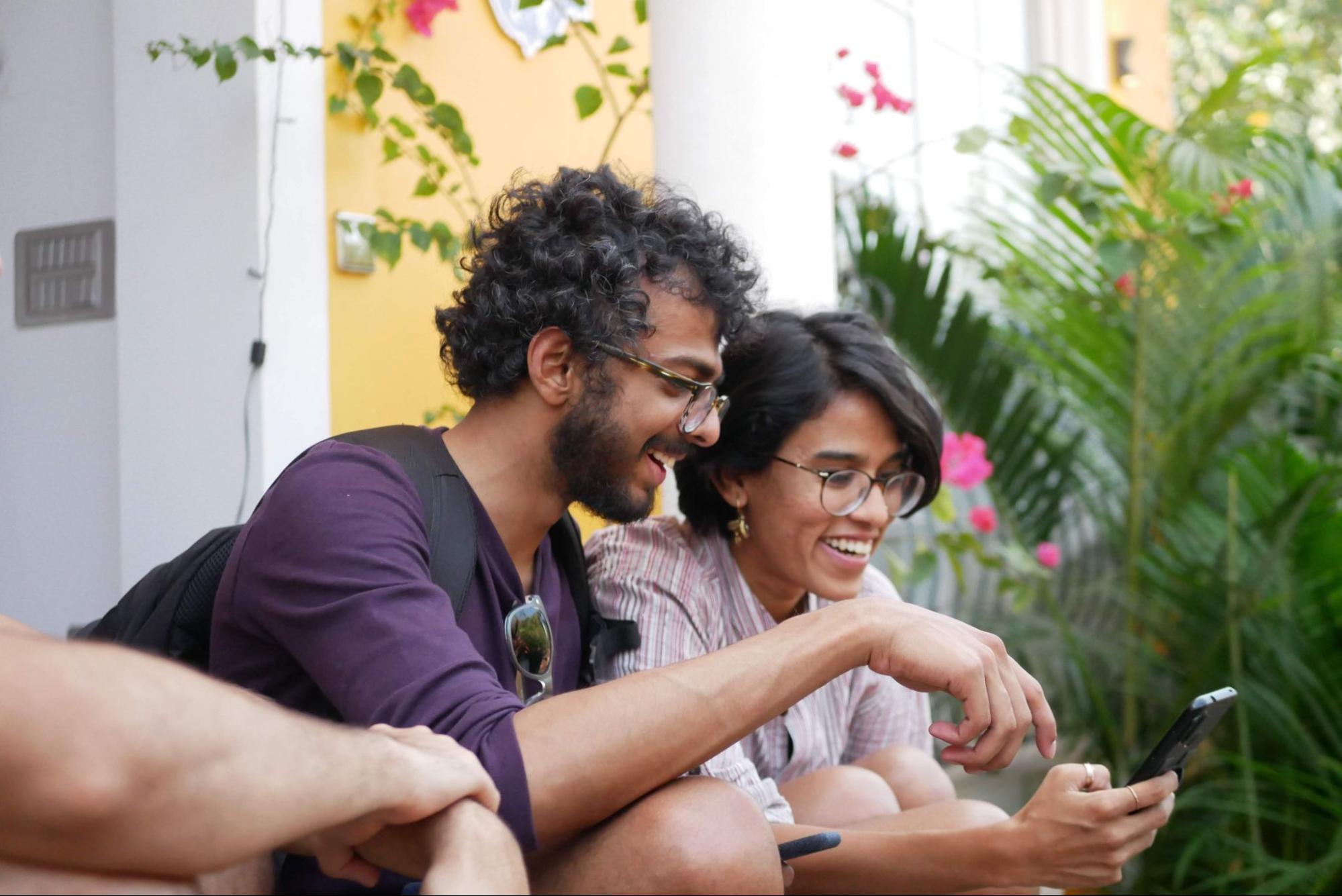 Imagem de duas pessoas sentadas, conversando, sorrindo e olhando para a tela de um celular que está à frente deles.