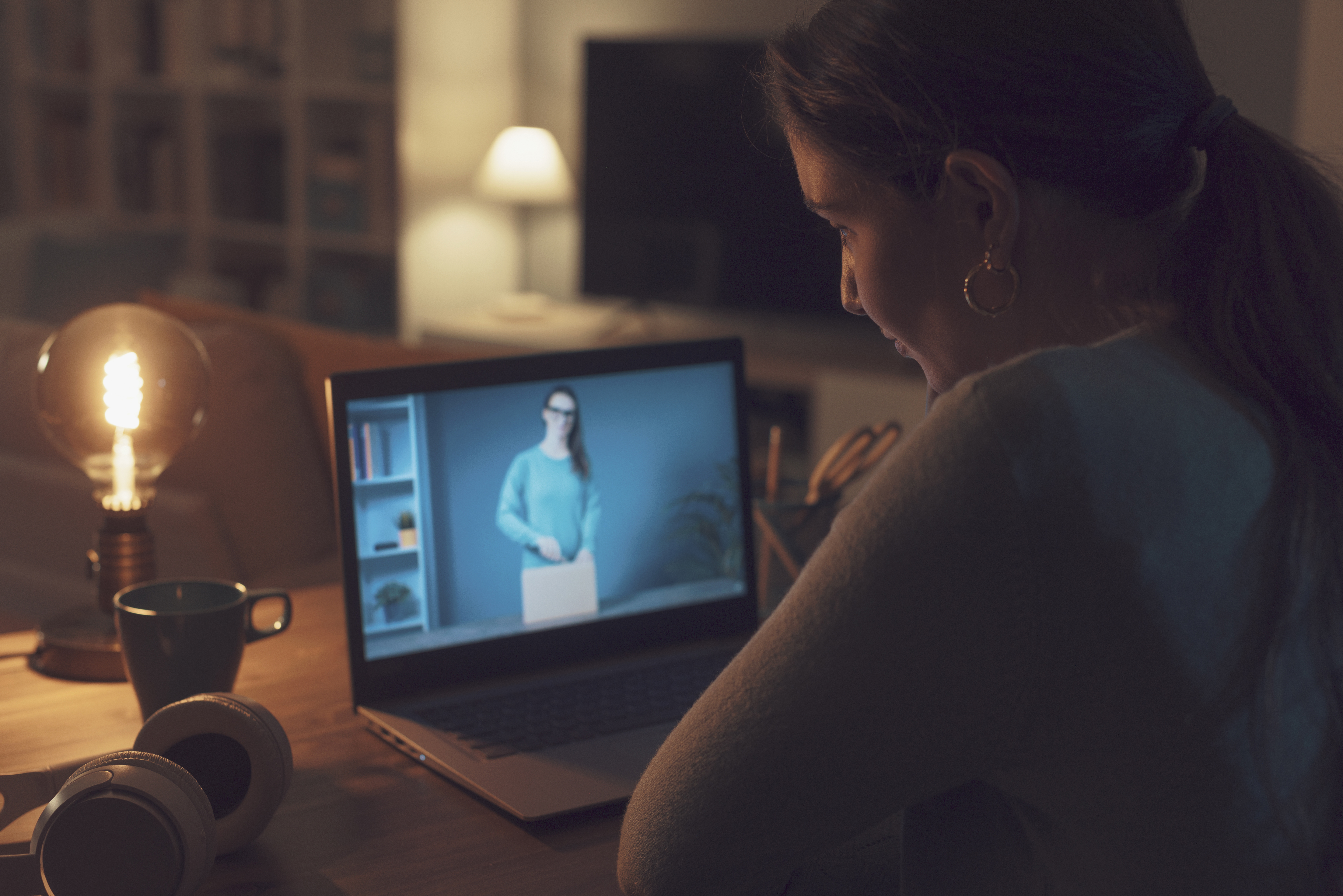 Fotografia de uma mulher sentada de frente para um notebook, na tela do notebook tem um video passando.