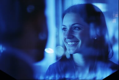 Uma mulher sorrindo enquanto conversa com outra pessoa, com um reflexo azul futurista no vidro, sugerindo uma interação digital ou tecnológica.